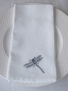 Black threaded embroidered dragonfly on white linen fabric napkin. Napkin is resting on traditional white dining plate.