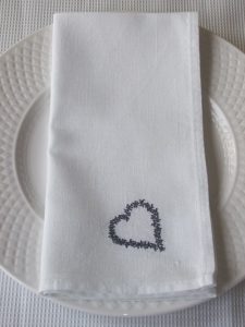 Black threaded embroidered heart on white linen fabric napkin. Napkin is resting on traditional white dining plate.
